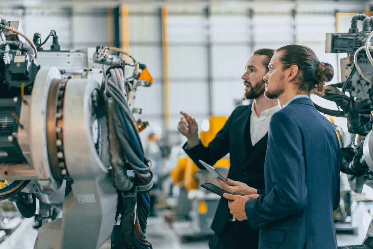 Two engineers discussing industrial machinery on a factory floor, with one holding a tablet.